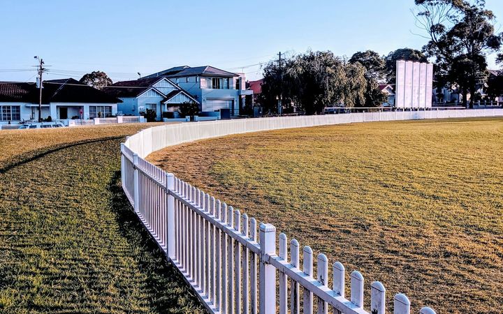 Long white picket fence curving around a grass field, houses behind